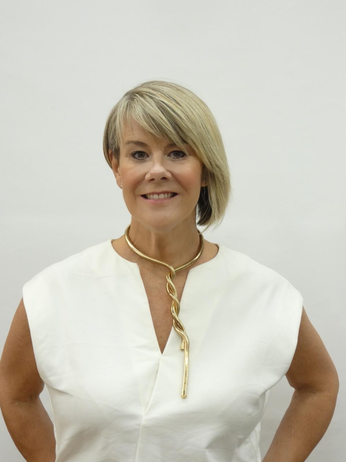 Elina James in a white top and a twisted gold statement necklace, smiling, hands on hips
